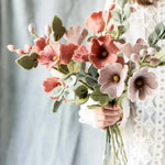 Person holding a bouquet of pink and dusty red flowers  and light green leaves against a neutral background