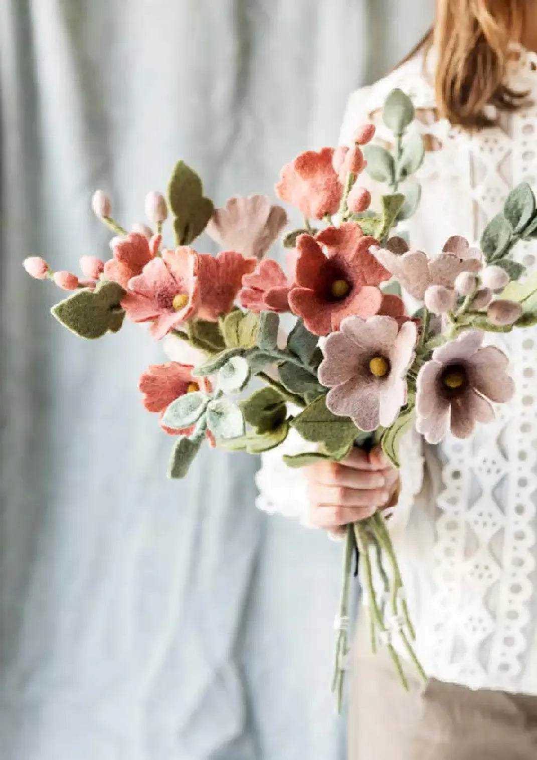 Person holding a bouquet of pink and dusty red flowers  and light green leaves against a neutral background