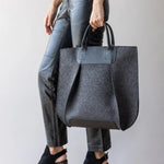 A person holds the graf lantz Frankie Tote, a large gray felt bag with black leather accents and handles. Dressed in black leather pants and heeled ankle boots, they display the totes structured elegance against a plain light background.