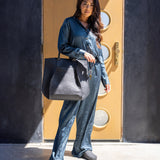 Dressed in a blue silk outfit, a person stands before a modern door with circular windows, holding a large gray Frankie Tote by graf lantz. Their slippers shuffle on the ground as sunlight casts shadows, highlighting their structured silhouette.