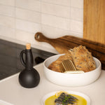 Bowl of bread and a plate of food on a kitchen counter with a black bottle and wooden cutting board.