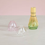 Three glass whisk stands in a pink, clear, and green base on a light gray surface with a pink background.