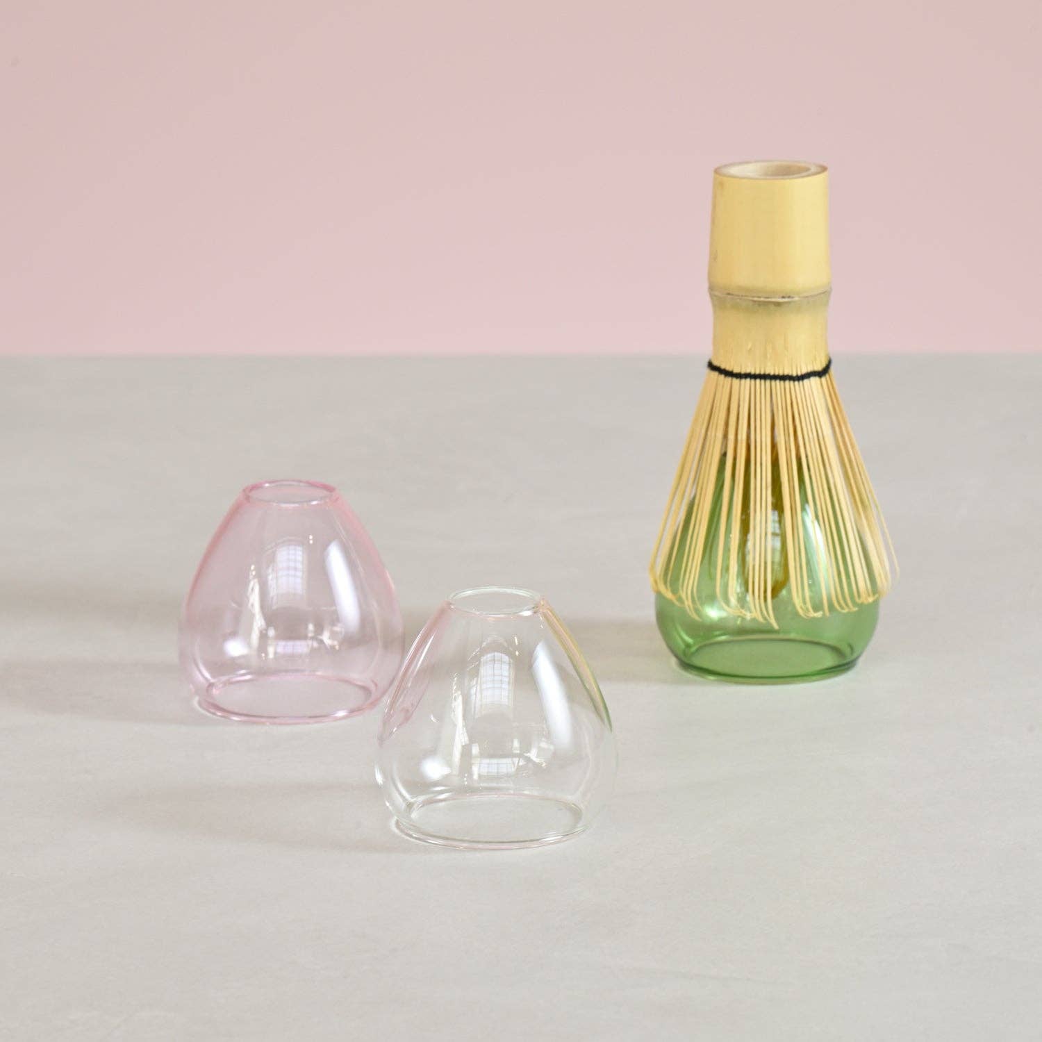 Three glass whisk stands in a pink, clear, and green base on a light gray surface with a pink background.