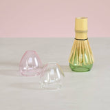 Three glass whisk stands in a pink, clear, and green base on a light gray surface with a pink background.