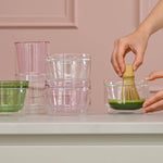 Person preparing a matcha using a whisk in a clear glass bowl on a countertop.