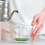 Person pouring water from a silver kettle into a clear glass bowl with matcha on a white background