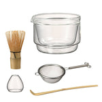 Matcha set including a whisk, glass bowl, strainer, whisk stand, and bamboo scoop on white background.