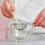 Person using a bamboo scoop and a strainer over a glass bowl on a white background