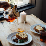 Person pouring tea into a glass on a wooden table with plates of cake and chocolate.