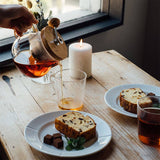 Person pouring tea into a glass on a wooden table with plates of cake and chocolate.