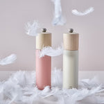 Two salt grinders in white and dark pink with wooden tops on a surface with feathers