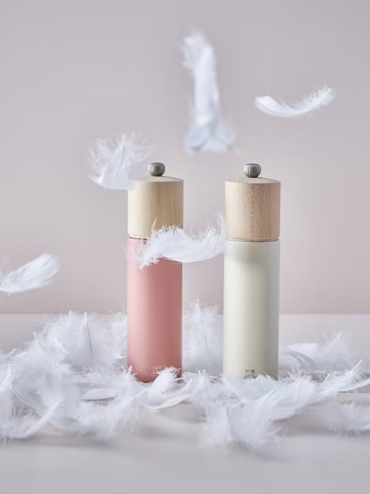 Two salt grinders in white and dark pink with wooden tops on a surface with feathers