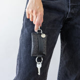 Standing woman holds charcoal Merino Wool Card Key Fob with two fingers in front of one leg in black jeans, front view, white background.
