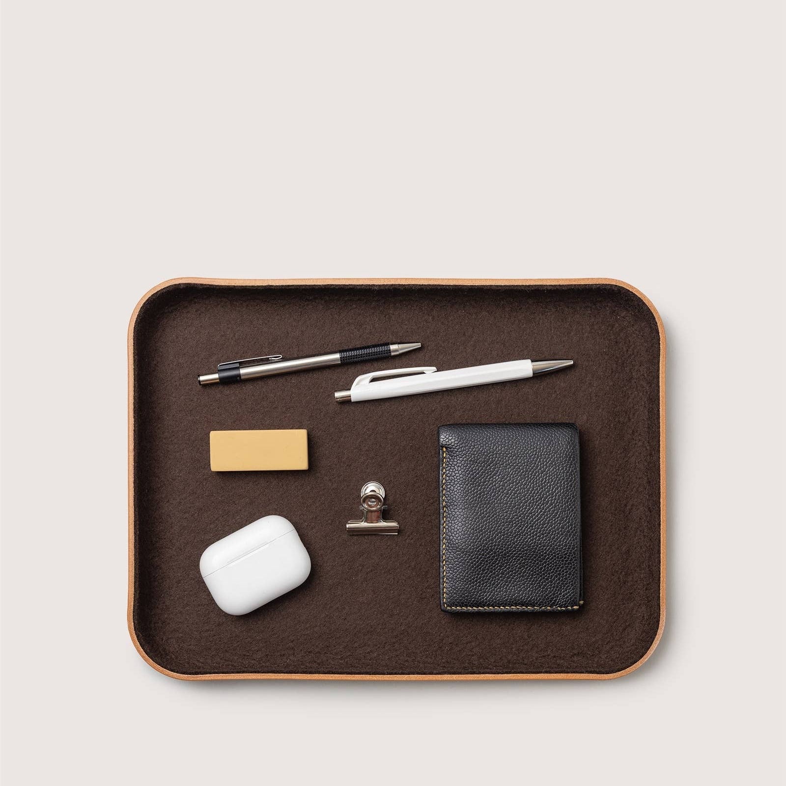 Brown tray with office supplies including a pen, airpods, clip, and wallet on a light gray background