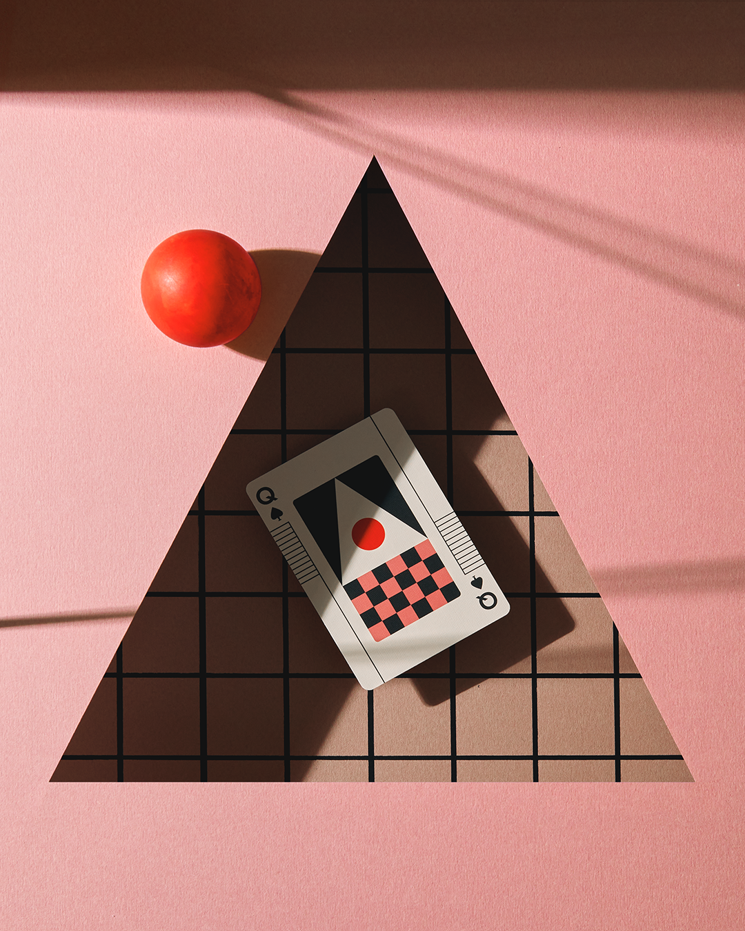 Card with a checkered design and red ball on a pink surface with a triangular shadow