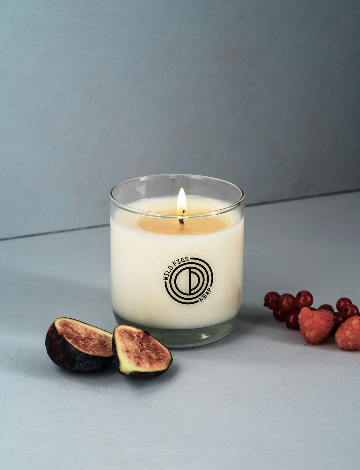 Candle in a glass jar with a logo, surrounded by figs and berries on a gray background