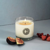 Candle in a glass jar with a logo, surrounded by figs and berries on a gray background