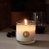 Candle in a glass jar with a logo on a wooden surface, surrounded by dark cherries.