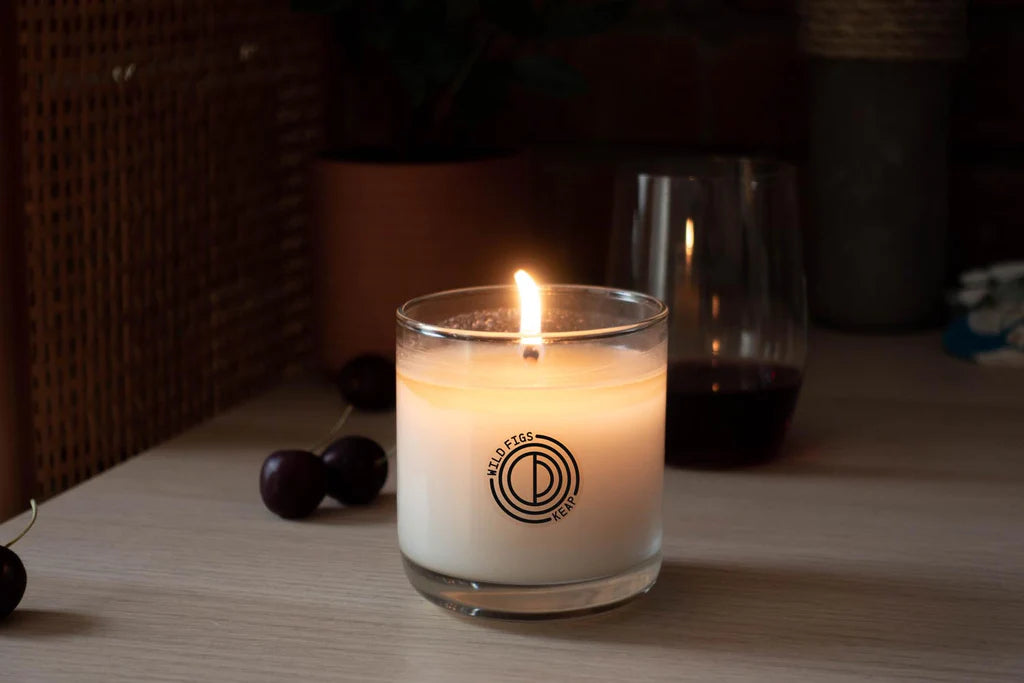 Candle in a glass jar with a logo on a wooden surface, surrounded by dark cherries.