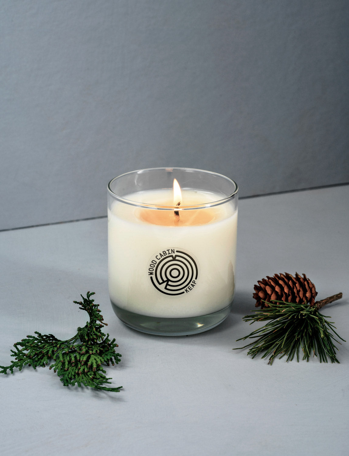Candle in a glass jar with a logo, surrounded by greenery on a gray background