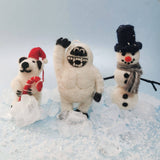 Three festive figurines: a polar bear, a creature in a snow suit, and a snowman, on a snowy background.