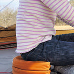 Torso of person in lavendar colored and white striped shirt and jeans sitting on a folded Snow Peak orange cushion. 