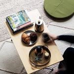 Snow Peak green cushion on a beige rug, next to a low table with tea and food. The arm of a person sitting outside thr frame holds a cup and is slightly blurred, suggesting motion. 