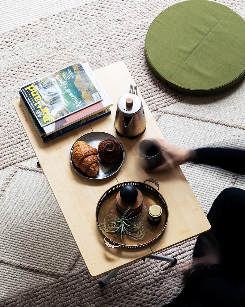 Snow Peak green cushion on a beige rug, next to a low table with tea and food. The arm of a person sitting outside thr frame holds a cup and is slightly blurred, suggesting motion. 