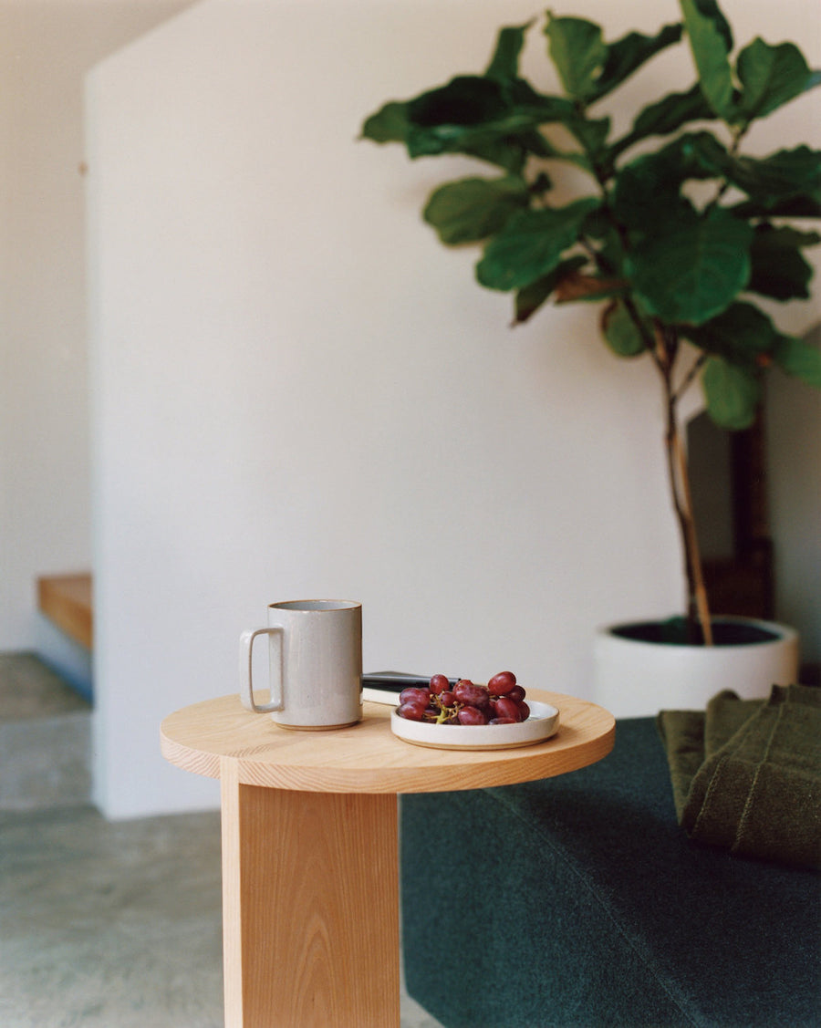 15 oz Hasami Porcelain mug in gloss grey is shown resting on a wood side table with a round top, next to a small white plate of purple grapes. There is a large ficus tree in the background against a white wall, and the corner of a dark green sofa is shown next to the side table.