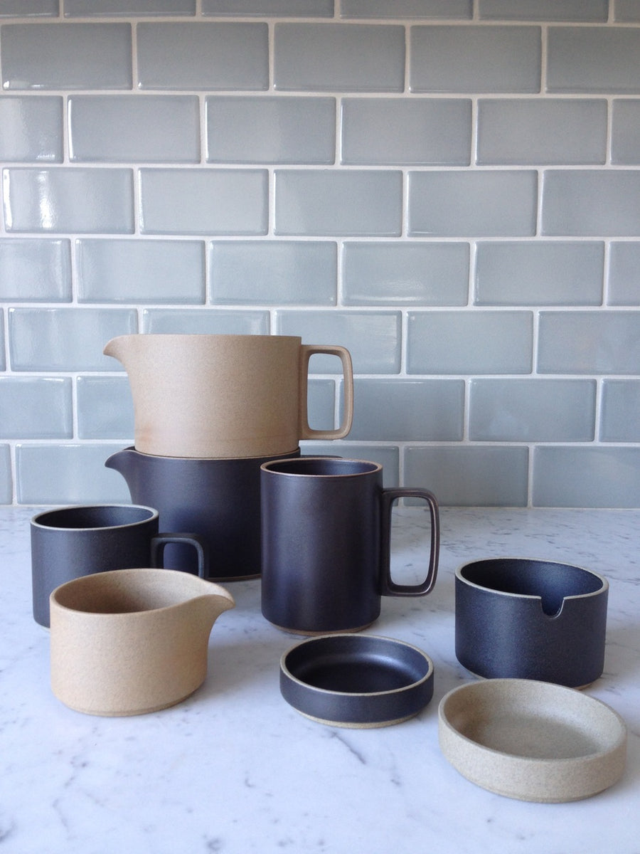 An assortment of Hasami Porcelain is shown on a marble counter top against a grey tile wall, from left to right: 11 oz black mug, natural creamer, black teapot with natural coffee dripper stacked on top, a black 15 oz mug, a black coaster, a natural coaster, and a black sugarpot with no lid on top.