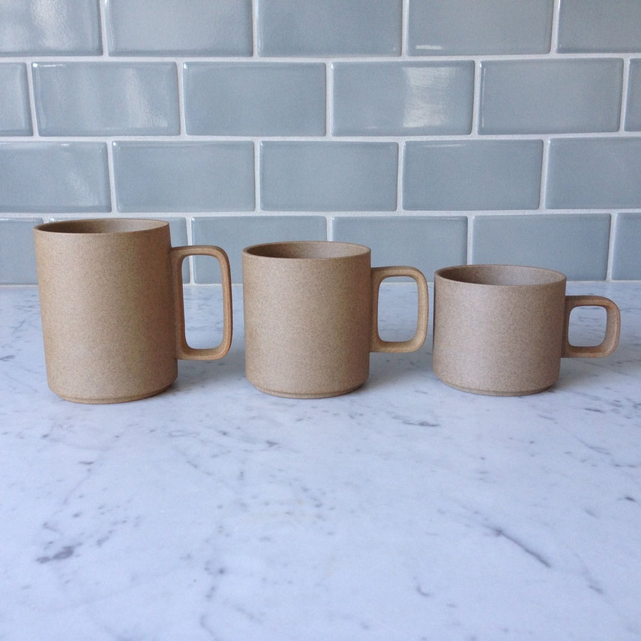 Three Hasami Porcelain mugs in Natural are arranged in a line in descending size order 15 oz, 13 oz, and 11 oz. The mugs are resting on a white marble countertop with a grey tile backsplash.