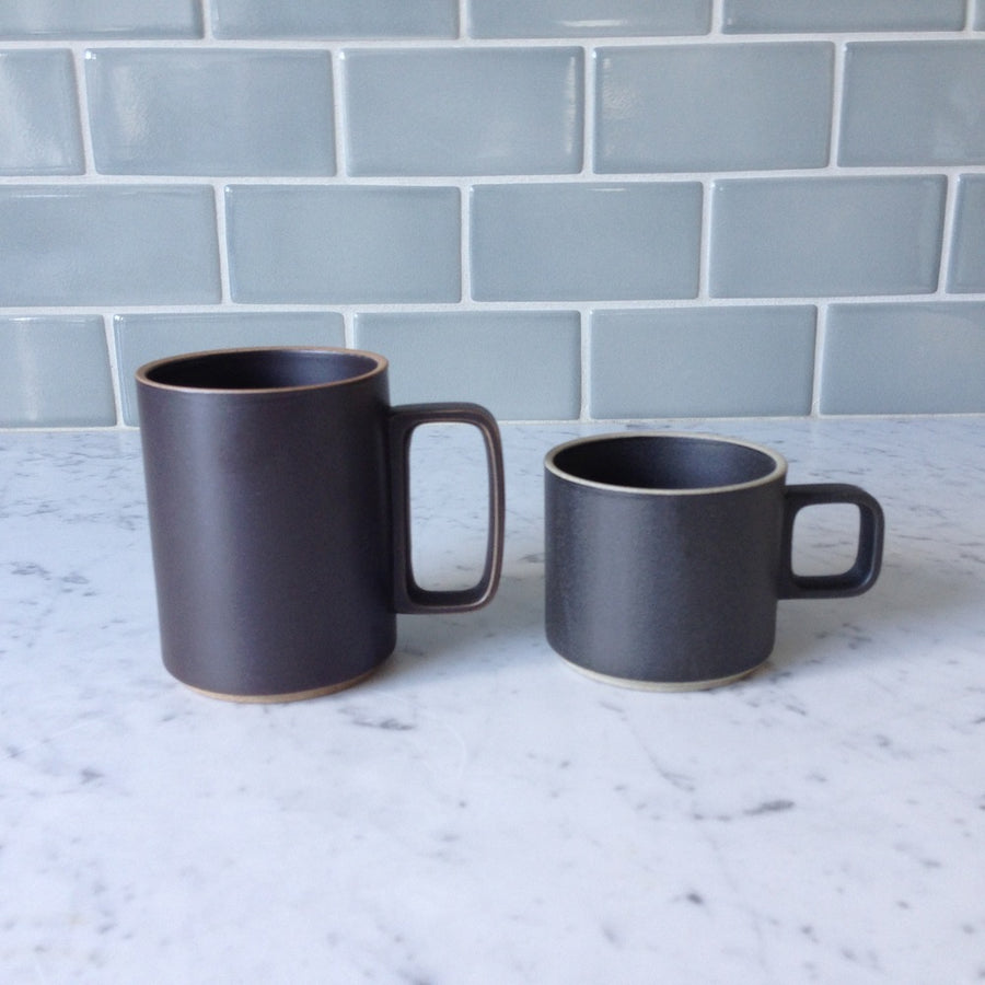 Two black Hasami Porcelain mugs are shown on a marble counter top against a grey tiled wall, 15 oz mug on the left and a 11 oz mug on the right.
