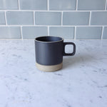 Hasami Porcelain 11 oz black mug resting on the natural coaster, sitting on a white marble counter top with a grey tile backsplash.