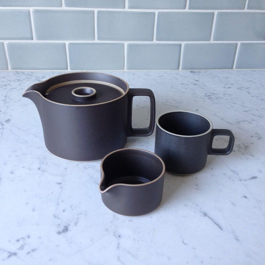 Black Hasami Porcelain teapot is shown resting a marble counter top, with a black 11 oz mug next to it, and a black creamer in the foreground.