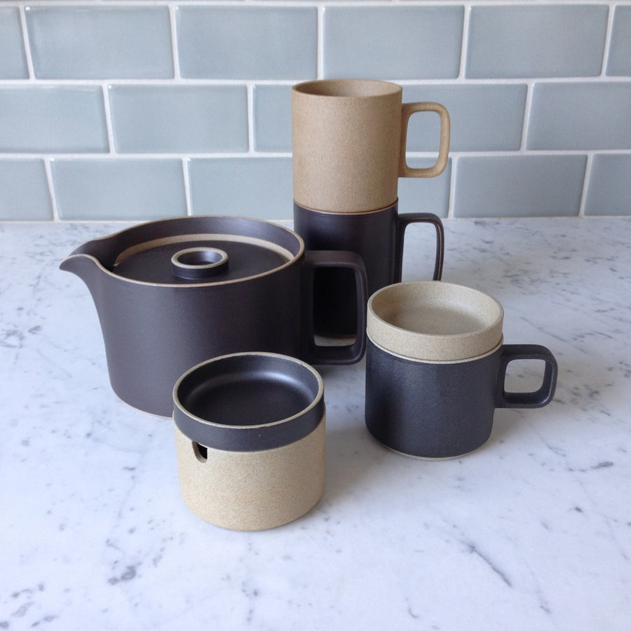 An assortment of Hasami Porcelain is shown a marble counter top: black teapot, black 11 oz mug with natural coaster, black 15 oz mug with a natural 13 oz mug stacked on top of it, and a natural creamer with a black lid.
