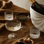 glass champagne coupes and low tumblers on brown velvet tablecloth next to bottle of champagne